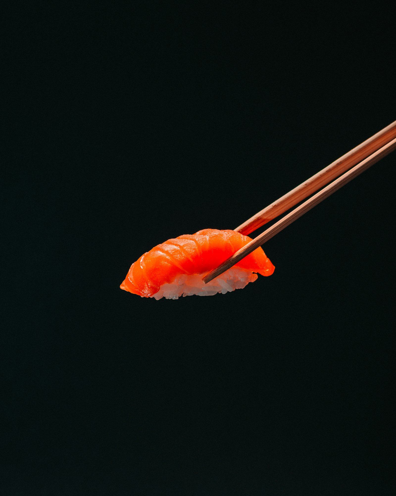 A single piece of salmon nigiri held in wooden chopsticks against a black background